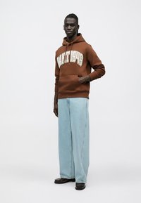 Sudadera marrón con capucha y logo "GANT 1949", bolsillo canguro, combinada con vaqueros azules de pierna ancha y zapatos negros. Fondo blanco liso.