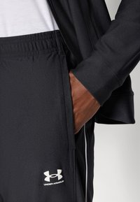 Pantalons de sport noirs en tissu texturé, avec une taille élastique, des poches latérales et un logo Under Armour blanc sur le bas de la jambe gauche.
