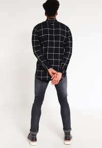 Uomo in piedi di spalle, vestito con una camicia a scacchi bianca e nera, jeans scuri arrotolati e sneakers scure con suole bianche.