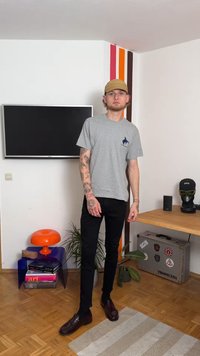 Szürke rövid ujjú póló egy kis kék logóval, fekete slim-fit nadrág és sötét cipő. A háttér színes falat és bútorokat ábrázol.