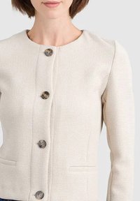 Giacca di colore crema con texture, collo rotondo, sei grandi bottoni frontali e due tasche laterali. Vestibilità sartoriale con dettagli alle spalle.