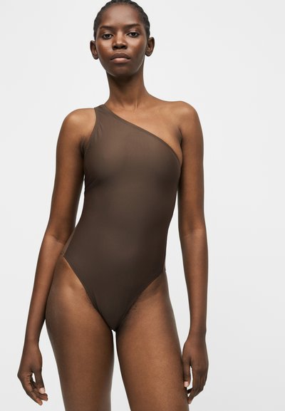 Γυναίκα που στέκεται με αυτοπεποίθηση φορώντας ένα μαγιό με ένα ώμο σε σκούρο καφέ χρώμα, μπροστά σε μια απλή ανοιχτόχρωμη επιφάνεια.
