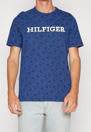 Mann trägt ein blau gemustertes, kurzärmeliges T-Shirt mit dem weißen Schriftzug "HILFIGER" auf der Brust und hellblaue Jeans.