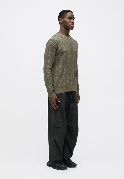 Ελαιοπράσινη πλεκτή μπλούζα με υφασμάτινες κάθετες ρίγες και σχέδιο λογότυπου. Συμπληρώνεται με χαλαρές μαύρες cargo παντελόνες και μαύρα αθλητικά παπούτσια.