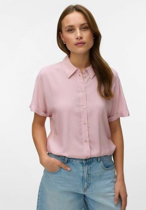 Kobieta o długich brązowych włosach, ubrana w krótki różowy sweter w paski zapinany na guziki oraz jasnoniebieskie dżinsy, stoi z jedną ręką w kieszeni.