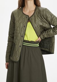 Chaqueta acolchada de color verde oliva con cierres a presión, llevada sobre una blusa amarilla brillante y una falda con cinturilla elástica de rayas verdes.