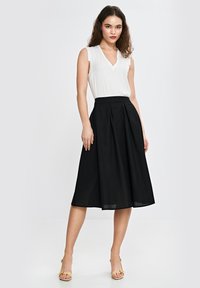 Biała bluzka bez rękawów z dekoltem w kształcie litery V, zestawiona z czarną, plisowaną spódnicą do kolan. Modelka w żółtych sandałach na obcasie.