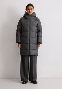 Langer, puffy Parka in Dunkelgrau, mit Kapuze, Reißverschluss-Taschen und gerippten Bündchen. Getragen über grauen weiten Hosen und schwarzen Schuhen.