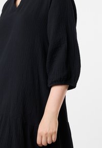 Musta mekko, jossa on teksturoitu kangas, V-pääntie, puffihihat ja kerroksellinen helma. Materiaali on pehmeästi ryppyinen.