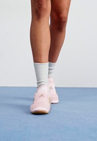 Rozā sporta apavi ar gludu tekstūru, ar zelta akcentiem un noapaļotu dizainu. Valkāti ar gaiši pelēkiem potīšu zeķēm uz zila virsmas.