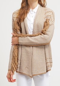 Beżowy i brązowy kardigan z plecionymi frędzlami, z teksturowanym wzorem i geometrycznymi akcentami. Otwarty fason, długie rękawy.