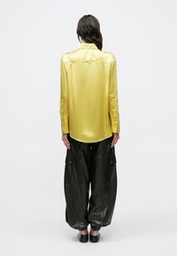 Cămașă galbenă din satin cu mâneci lungi, tiv curbat și manșete cu nasturi, combinată cu pantaloni largi negri cargo cu buzunare laterale.