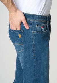 Jeans in denim di un blu medio con un taschino anteriore dotato di un logo ricamato giallo e un etichetta rossa sulla vita.