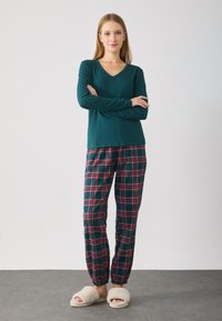 Grøn langærmet V-hals top parret med ternede pyjamabukser i mørk grøn, rød og hvid. Bløde beige indersåler med en plysséagtig tekstur.