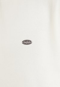 Petit badge métallique ovale avec le mot en cursive "Smilodox" attaché à un fond en tissu blanc uni.