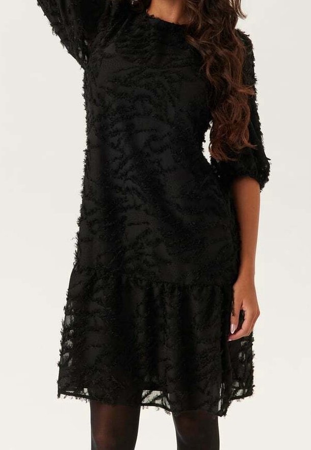 Femme portant une robe noire texturée longueur genou avec manches trois-quarts et un ourlet à volants, associée à des collants noirs.
