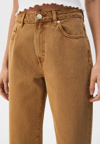 Gros plan d'une personne portant un jean marron taille haute et un top blanc court à ourlet effiloché, les mains détendues le long du corps.