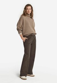 Beige gestrickter Rollkragenpullover, kürzere Passform. Braune weite Hose mit Taschen. Weiße Sneakers mit hellbraunen Akzenten.