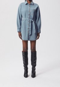 Vestido de camisa de mezclilla en azul claro, con cierre de botones en la parte delantera, mangas largas y cinturón de la misma tela para atar. Combinado con botas de cuero negro hasta la rodilla.