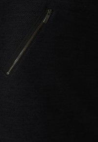 Černá textilie s kapsou na zip a kovovým kováním. Materiál má texturovaný povrch, který dodává designu hloubku.