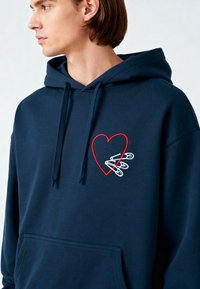 Felpa blu navy realizzata in morbido tessuto, con un cuore rosso e spilli di sicurezza ricamati sul petto. Include una tasca anteriore e un cappuccio con cordino.
