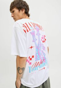 Balta T-kreklā ar krāsainu grafisko dizainu, kurā attēloti šaha figūras un uzraksts "Čempioni" sarkanā un zilā krāsā, ar šaha laukuma akcentiem.