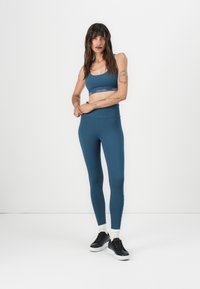Blå atletisk crop top med et merket bånd, parret med matchende høyhalsede leggings. Stylet med svarte sportsko og hvite ankelsokker.
