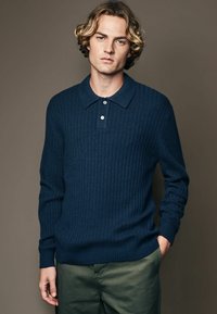Marineblauer gerippter Strick-Polo-Pullover mit klassischem Kragen und Knopfleiste mit drei Knöpfen. Lange Ärmel besitzen rippierte Bündchen. Getragen mit grünen Hosen.