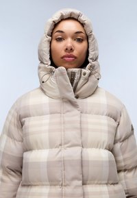 Puffjacka i dämpad beige med rutmönster, utrustad med huva, snördetaljer och en vadderad design för värme och komfort.