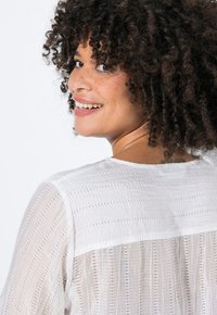 Blusa blanca con textura, con patrones de rayas horizontales y verticales, que presenta un escote redondo y tejido transparente.