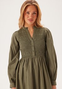 Robe longue à manches longues de couleur vert olive avec un corsage smocké, un devant à boutons et une jupe volée. Tissu doux avec un léger éclat et une finition texturée.