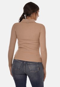 Maglione leggero beige chiaro a costine, aderente, con maniche lunghe e colletto. Presenta un piccolo bottone decorativo sul retro. Indossato con jeans in denim scuro.