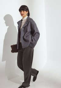 Pelēks vilnas maisījuma žakets ar izgrieztu apkakli, pogu aizdari un sānu kabatām, savienots ar krēmkrāsas turtleneku un tumšiem platās kājas biksēm.