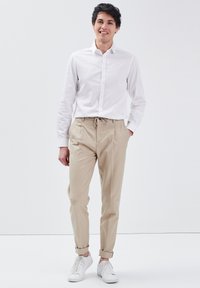 Witte blouse met knopen, beige tapered chinos met trekkoord in de taille, opgerolde mouwen en witte sneakers. Eenvoudige, casual outfit op een effen achtergrond.