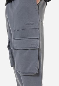 Närbild av grå cargobyxor med stora sidofickor och "PEGADOR" broderat ovanför en ficka. Lätt tygstruktur synlig.