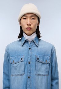 Lysblå denimskjorte med to brystlommer, kontrasterende stikninger og mørke knapper. Under skjorten er der en hvid mock turtleneck. Hvid hue bæres.