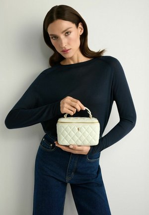 Kobieta o brązowych włosach, ubrana w czarną bluzkę z długim rękawem i niebieskie dżinsy, trzyma małą białą pikowaną torebkę z górnym uchwytem i zamkiem.