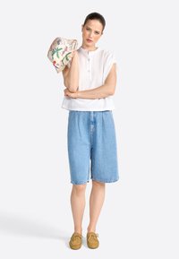 Chemise blanche à manches courtes à boutons, bermuda en denim bleu clair et chaussures en suède beige. Le mannequin tient un chapeau à motifs colorés.