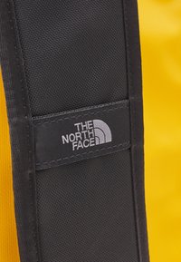 Black fabric strap with stitching and a logo patch, set against a bright yellow background, featuring a textured surface.