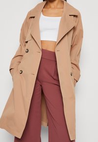 Beżowy płaszcz trencz z klapami z wycięciem, dwurzędowym przodem i kieszeniami bocznymi, noszony na białym krótki topie i bordowych spodniach.