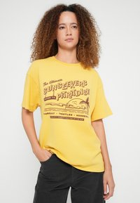 Kollane puuvillane t-särk lõdva lõikega, millel on pruuni teksti ja kujundusega graafiline trükk, kujutades laineid ja palmipuid.