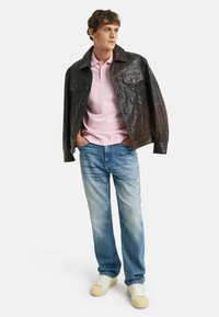 Giacca di pelle marrone, polo rosa chiaro, jeans blu scoloriti e sneakers bianche. Modello in piedi con le mani nelle tasche. Sfondo pulito e minimalista.