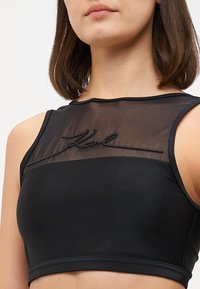 Svart crop topp med en gjennomsiktig mesh-overdel. Fronten har et brodert logo, glatt stoff og en tettsittende design.