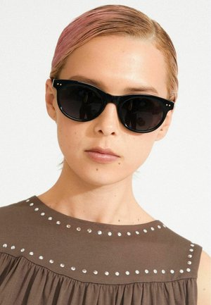 Melnas kaķa acu saulesbrilles ar tumšiem stikliem, ar maziem zelta akcentiem rāmī. Modelis valkā brūnu augšu ar sudraba kniedēm.