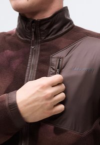 Χέρι ατόμου που κλείνει με φερμουάρ την τσέπη στο στήθος ενός καφέ μπουφάν Salomon με υφασμάτινα πάνελ με υφή και λεία επιφάνεια.