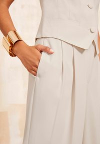 Beige tailored garment with pleats, featuring a sleek, structured design. A shiny gold cuff bracelet adorns the wrist, complementing the outfit.