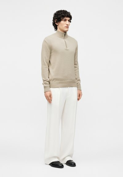 Noor mees seisab, kannab beeži lukuga kaelusega kampsunit, laiu valgeid pükse ja musti kingi, tavalise heleda tausta ees.