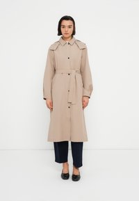 Manteau long beige avec une taille ceinturée, des boutons à l'avant et des épaulettes. Porté avec un pantalon foncé et des chaussures noires, présentant une texture lisse.