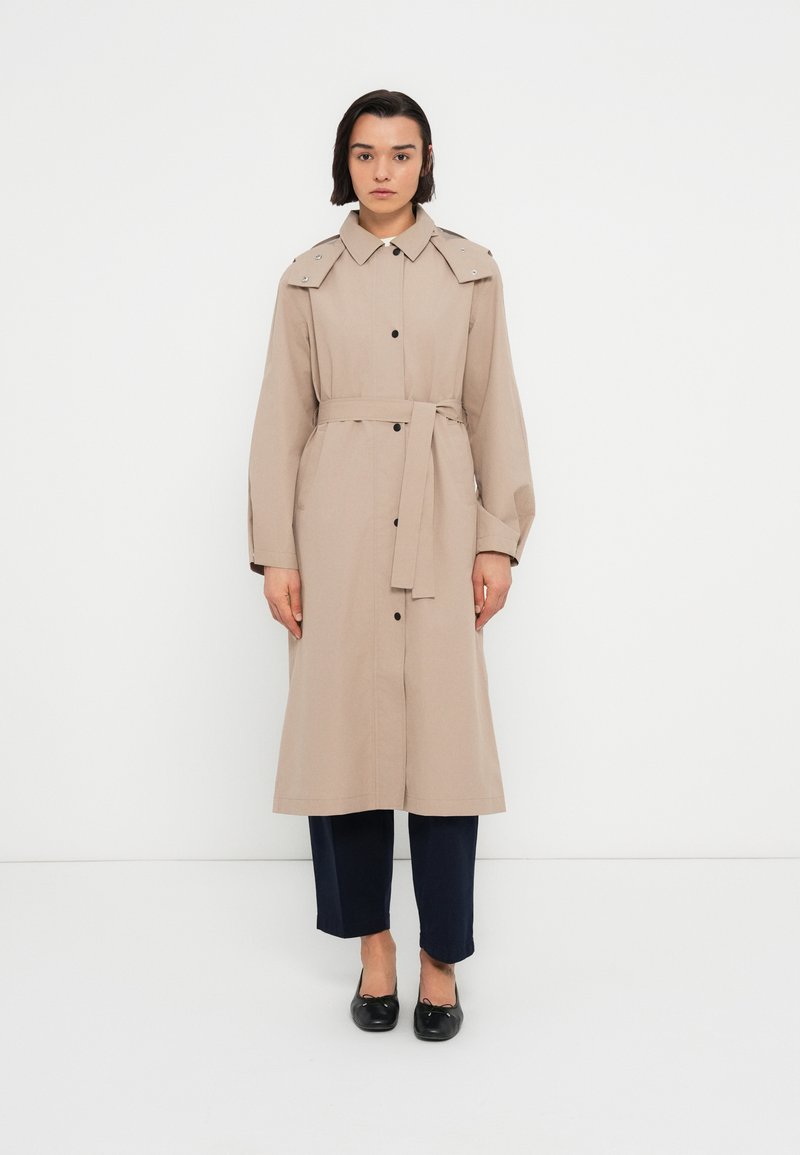 Manteau long beige avec une taille ceinturée, des boutons à l'avant et des épaulettes. Porté avec un pantalon foncé et des chaussures noires, présentant une texture lisse.