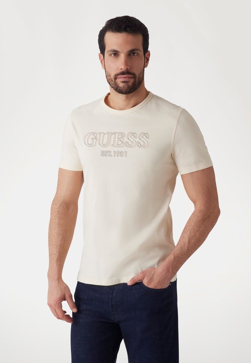 Camiseta de manga corta en color crema con el logo bordado "GUESS EST. 1981". Material de algodón suave, escote redondo y diseño ajustado.
